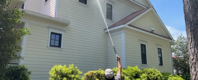 Person pressure washing the exterior of a large, light yellow house surrounded by greenery, representing pressure washing Wilmington NC.