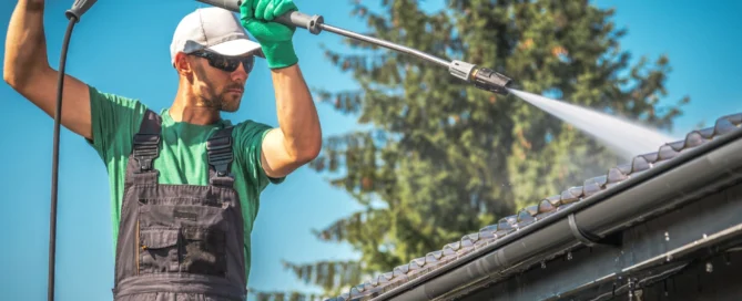 Person wearing work gear pressure washes a roof, with clear sky and trees in background. Related to pressure washing Wilmington NC.