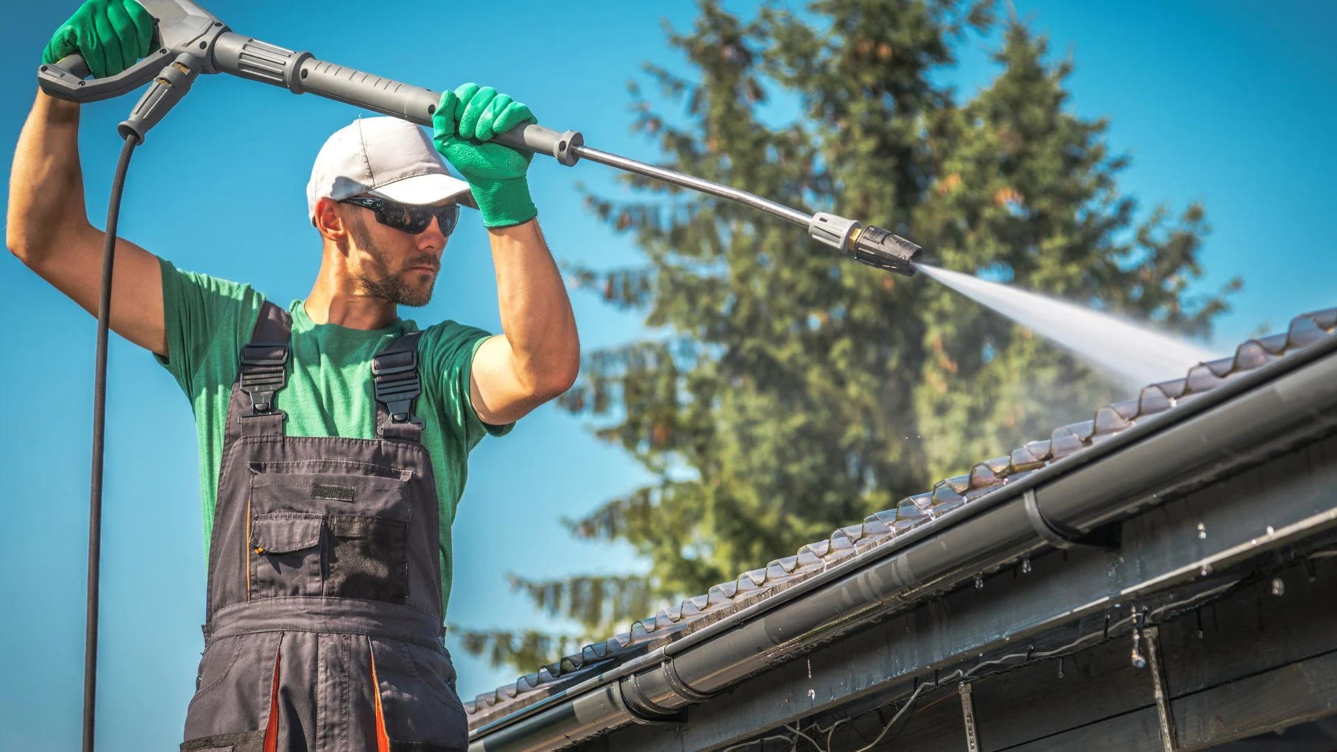Person wearing work gear pressure washes a roof, with clear sky and trees in background. Related to pressure washing Wilmington NC.