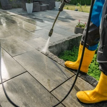 Person in yellow boots uses a pressure washer on a patio. Clean patio stones gleam under the sun. Pressure washing Wilmington NC.