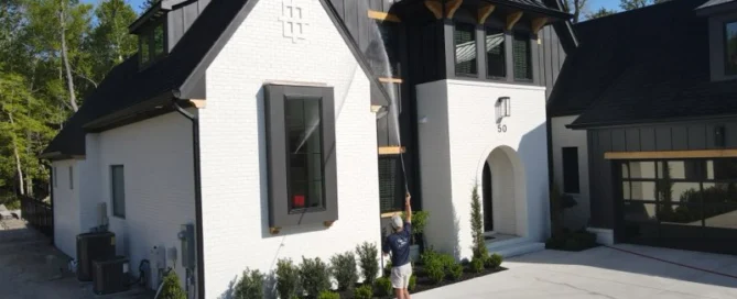 Person pressure washing a modern white house with black trim in Wilmington, NC, surrounded by trees and a clean driveway.