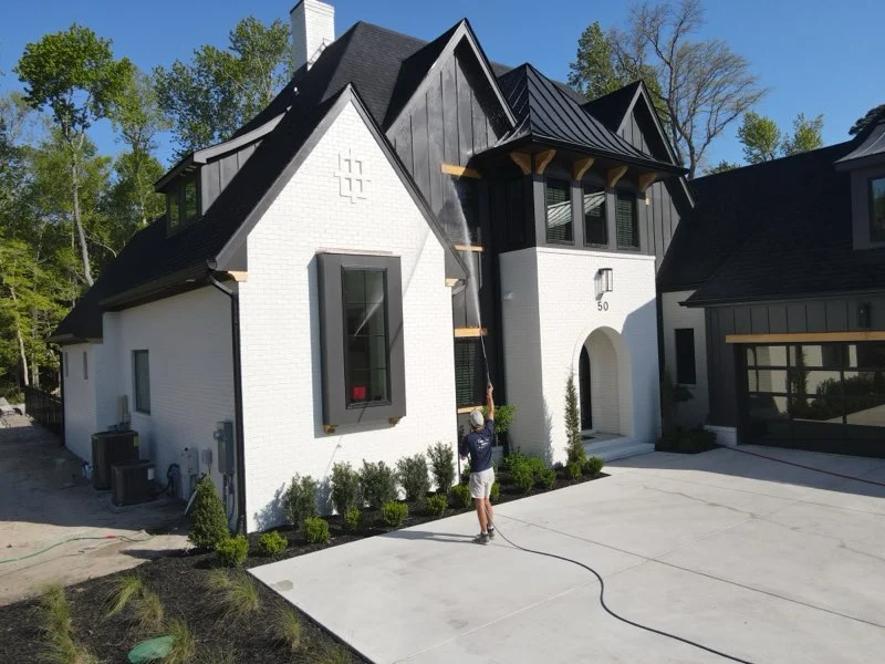Person pressure washing a modern white house with black trim in Wilmington, NC, surrounded by trees and a clean driveway.