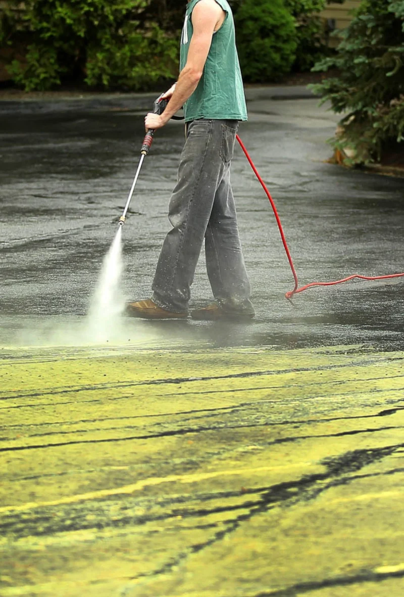 Person pressure washing a driveway, removing dirt and stains. Red hose trails on wet surface, greenery in background. Pressure washing Wilmington NC.