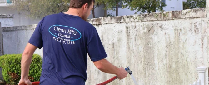 Worker in blue shirt pressure washing a stained wall near fencing, showcasing pressure washing Wilmington NC service.