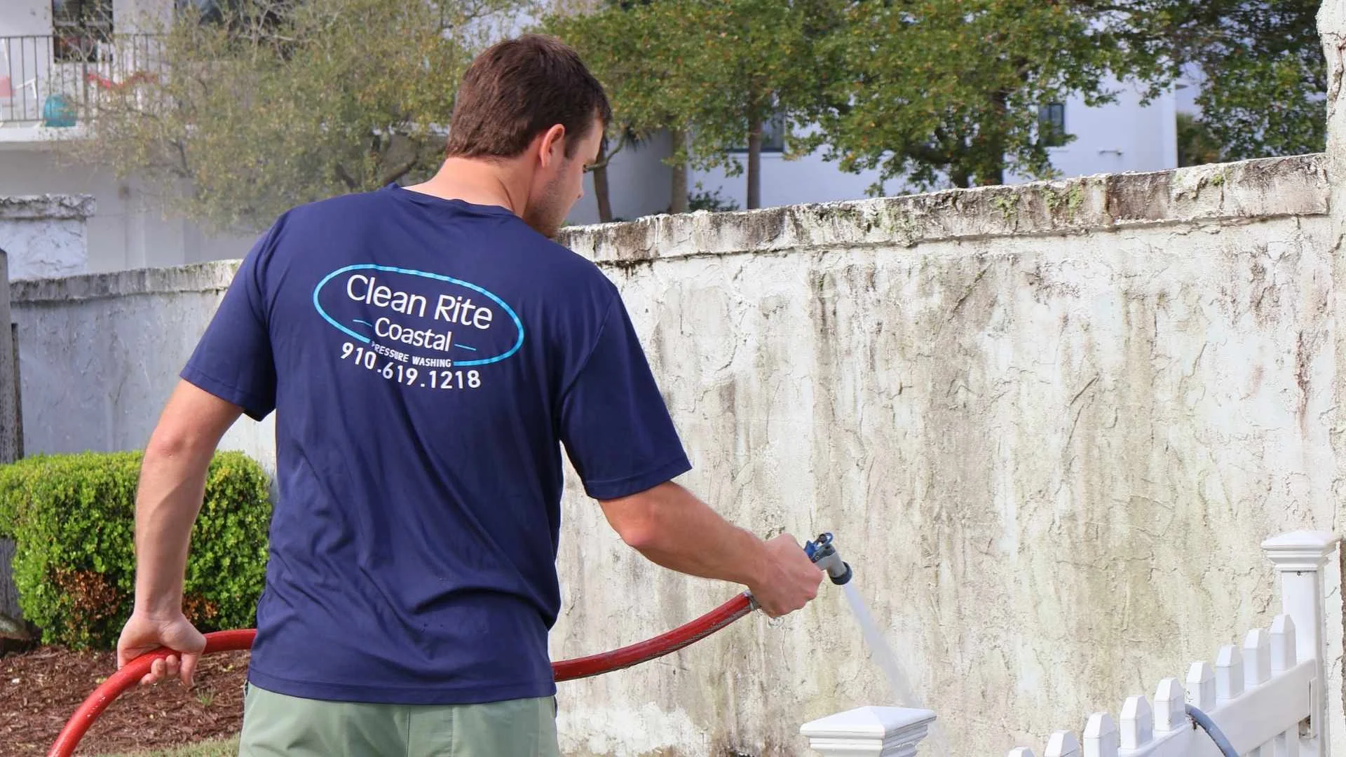 Worker in blue shirt pressure washing a stained wall near fencing, showcasing pressure washing Wilmington NC service.