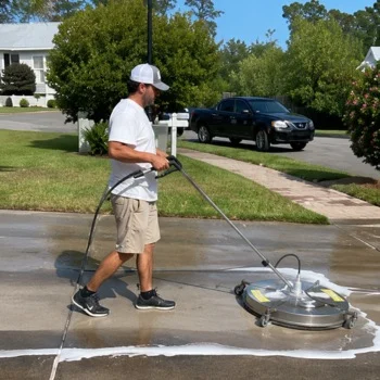 Sidewalk and Curb Pressure Washing HOAs Person using a large surface cleaner for pressure washing a driveway. Green lawn and parked car visible. Sunny day in Wilmington, NC.