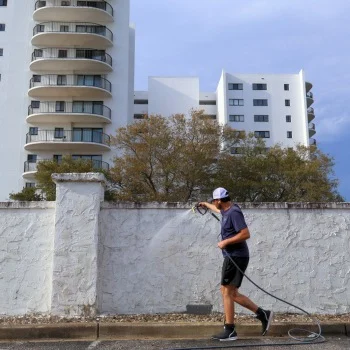 Person using a hose to pressure wash a white wall by a modern apartment building; related to pressure washing Wilmington NC.