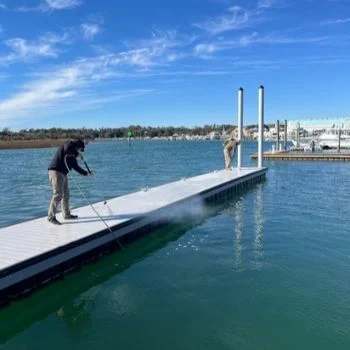 What Surfaces Should Be Soft Washed Instead of Pressure Washed SMALL Two people pressure washing a dock on a sunny day in Wilmington, NC, with clear blue skies and calm water.